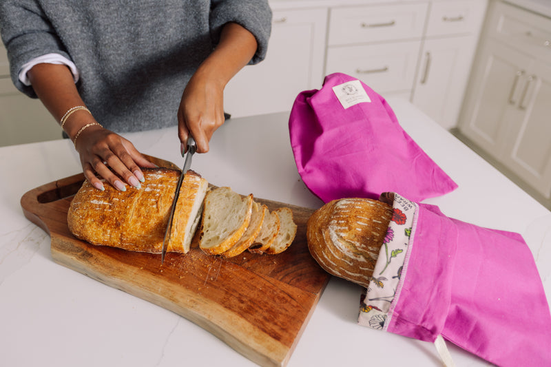 Person slicing bread on a wooden cutting board with Meadow Magic Bread Bags and loaf of bread in the background.